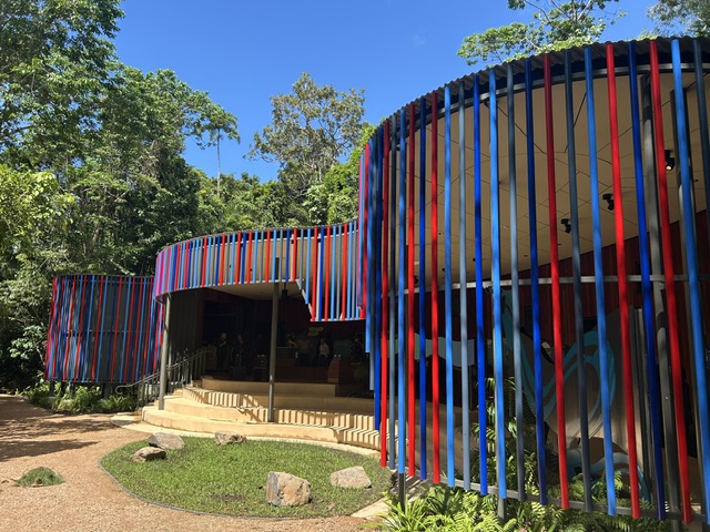 Colourful building with curved walls covered in vertical red and blue poles, surrounded by trees and greenery, with four steps leading to the entrance of the building.  